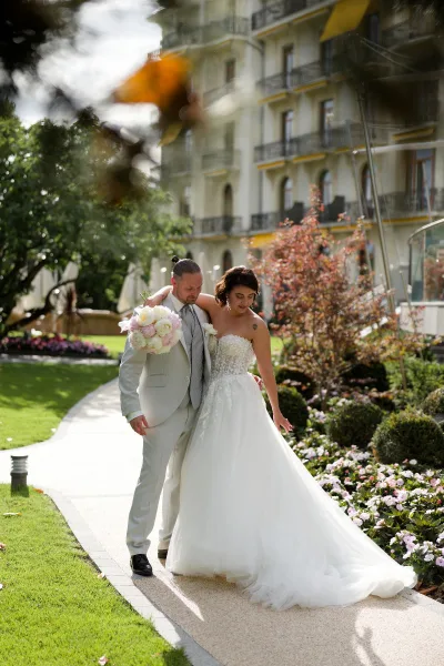 Couple portrait of the bride and groom walking along a garden walkway, her peony bridal bouquet bright as flower beds and shrubs frame the scene.