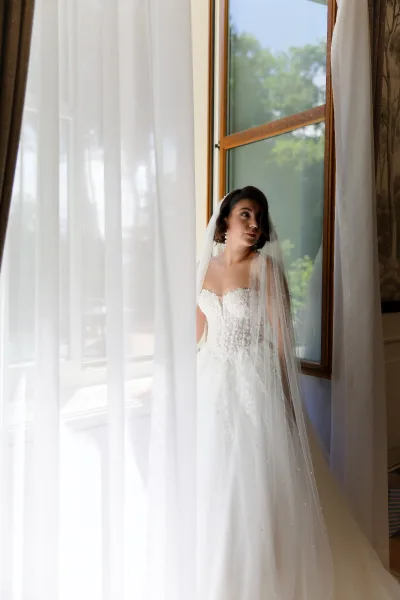 Bridal portrait of a bride by the window in a lace wedding dress, bathed in soft natural light through sheer curtains with trees beyond.