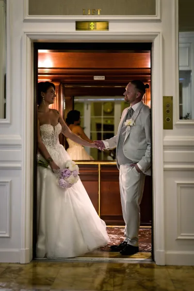 Couple portrait of a bride and groom holding hands in a hotel elevator, her pastel bridal bouquet bright against the wood paneling and marble floor.