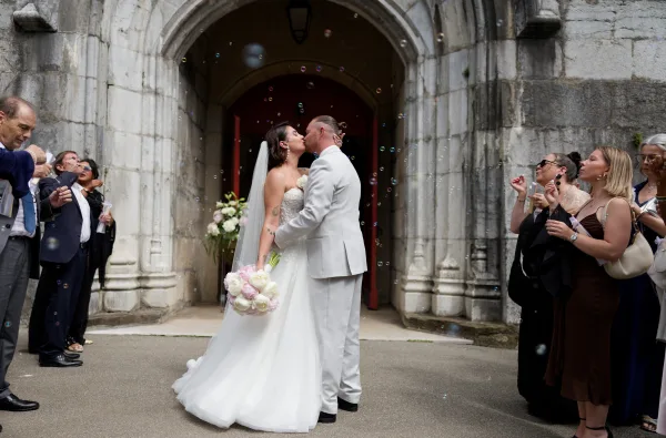 Wedding bubble exit as the couple share a ceremony exit kiss under a stone archway by red church doors, her veil trailing softly.