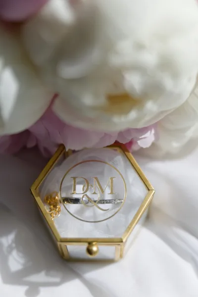 Wedding ring box, a glass hexagon ring box nestled on white fabric, framed by scattered peony petals.