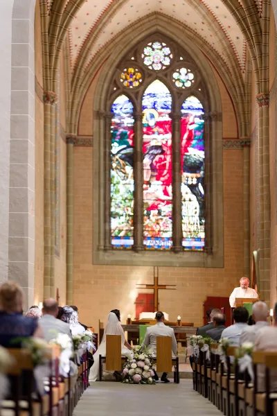 Ceremony moment at a church wedding ceremony with the couple at the altar before a cross, beneath a stained glass window, amid stone walls and pews.