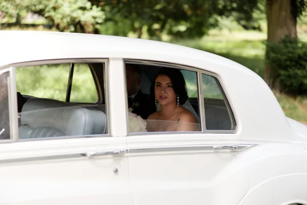 Bride in car gazes from a classic white wedding car, holding a bouquet, framed by leafy park trees and greenery.