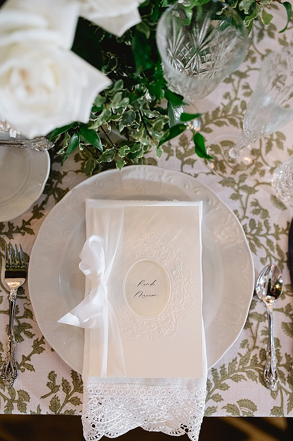 Wedding place setting with wedding menu card tied in a white ribbon atop lace napkin, white plates, silver flatware, and crystal glassware with roses