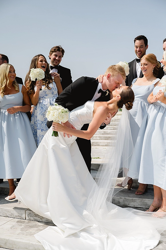 Wedding kiss portrait of bride and groom in a dip kiss wedding photo as bridesmaids cheer on outdoor steps under open sky