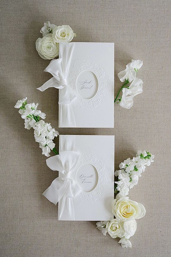 Wedding invitation suite with embossed wedding invitations tied in a white ribbon bow, surrounded by ranunculus and sweet pea on linen backdrop
