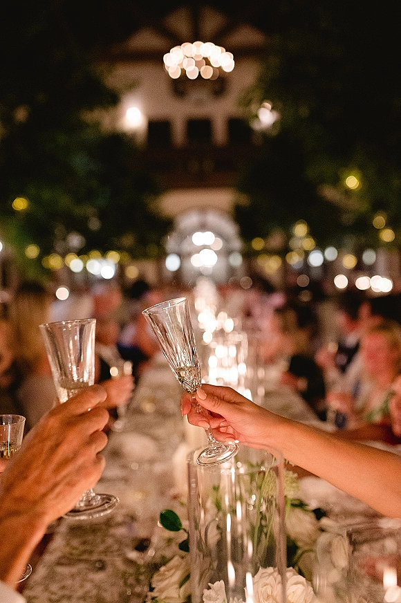 Wedding toast with champagne toast photo as guests clink flutes over candlelit banquet table with white florals and string lights at night