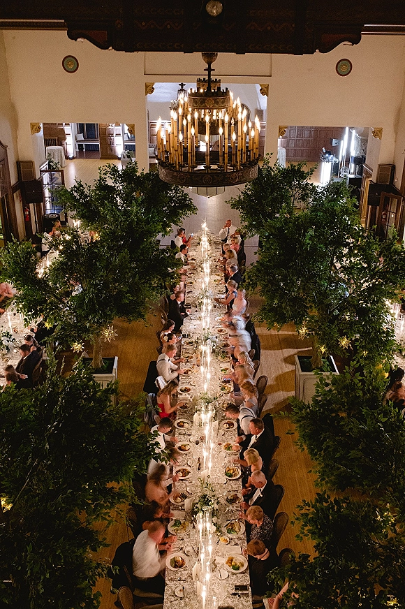 Wedding reception tablescape with long banquet table setup, taper candles, white florals, greenery garlands, and string lights in an indoor hall