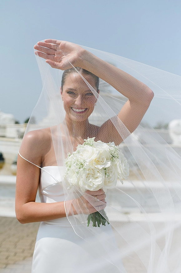 Bridal portrait of a smiling bride holding bouquet of white roses under a wedding veil at a waterfront marina with boats and blue sky