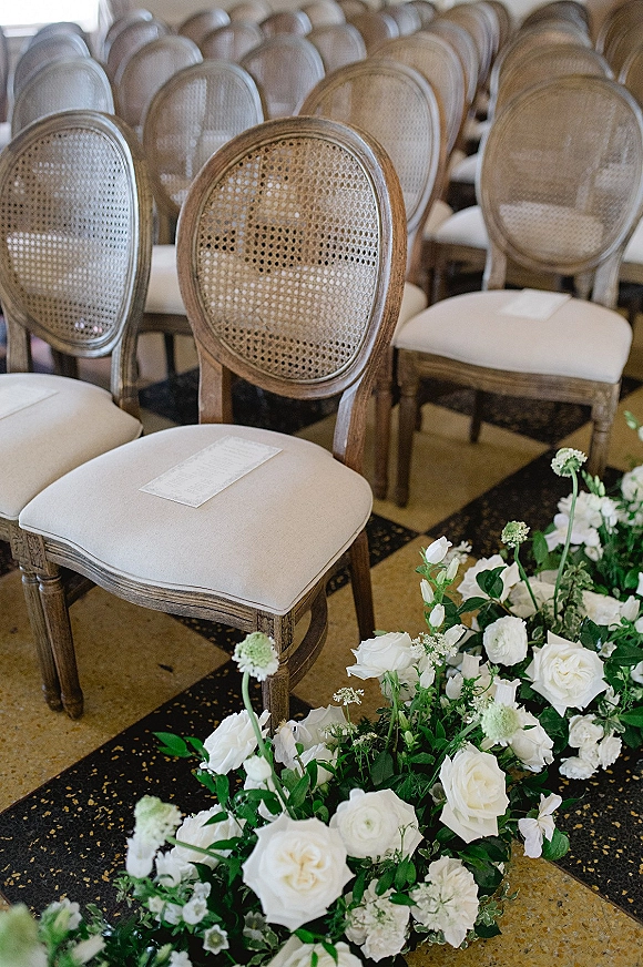 Ceremony seating with wedding chair rows of cane back chairs, cushioned seats and programs, plus white rose and greenery aisle florals indoors