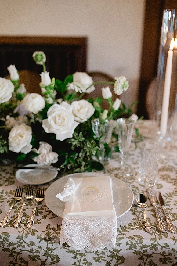 Reception tablescape with wedding place setting, white rose centerpiece and greenery, lace napkin, ribbon-tied menu card, and taper candle in hurricane glass