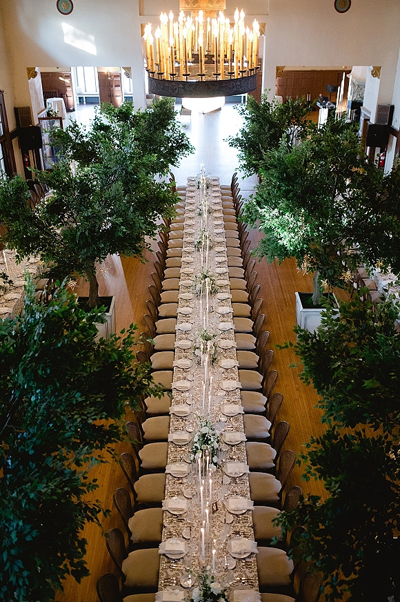 Reception tablescape with a long banquet table set with candles, white-green florals, greenery runner, and chandelier-lit indoor hall