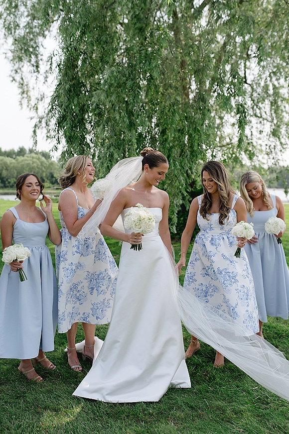 Bride with bridesmaids walking on a green lawn, carrying white rose bouquets as her long veil blows near a lakeside tree under overcast sky