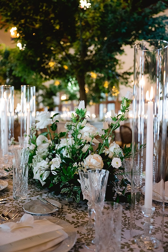 Reception tablescape with wedding table centerpiece of white florals and greenery garland, taper candles, and string lights in a softly lit room
