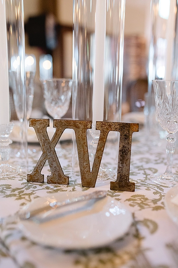 Table number decor featuring a metal table number beside tall glass cylinder vases, crystal goblets, and patterned tablecloth at an indoor reception table