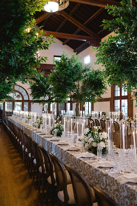 Reception tablescape with a long banquet table wedding setup featuring white floral centerpieces, candlelight, and greenery under string lights and wood beams