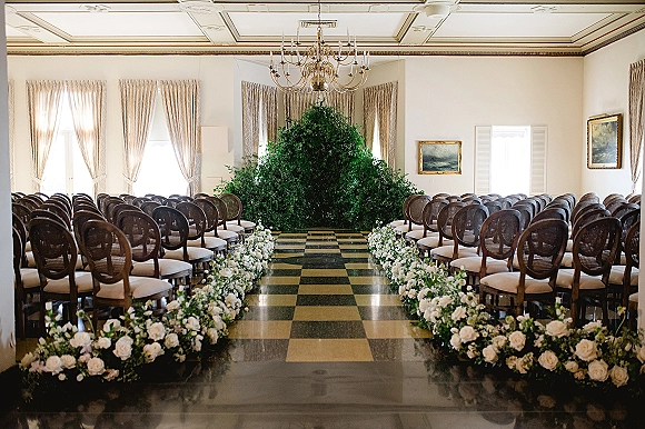 Ceremony setup for indoor wedding ceremony with white rose aisle flowers and greenery, chandelier overhead in a ballroom with checkered floor