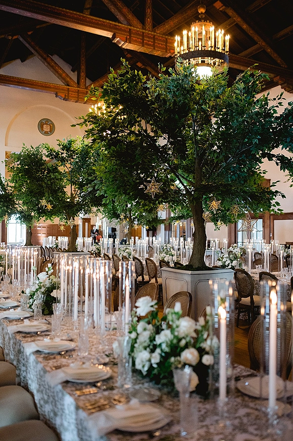 Reception tablescape with a long banquet table, white floral and greenery centerpieces, taper candles, and star ornaments under a chandelier