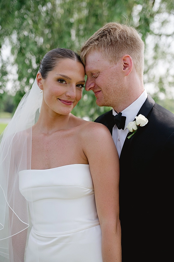 Couple portrait of bride in strapless wedding dress and veil as groom in tuxedo nuzzles her forehead on a green outdoor lawn backdrop