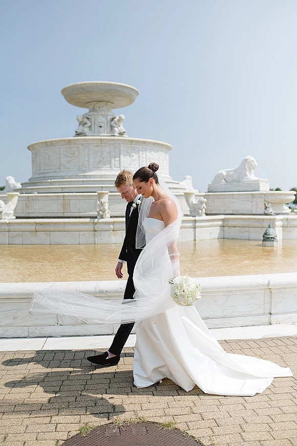 Couple portrait of bride and groom walking, her veil blowing as she holds a white bouquet beside a stone fountain and reflecting pool