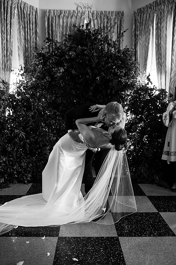 Wedding kiss portrait of bride and groom in a dip kiss wedding photo, long veil flowing, chandelier overhead and greenery backdrop behind