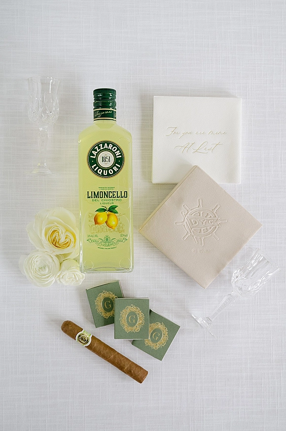 Wedding flat lay wedding invitation flat lay with embossed invitation box, white roses, limoncello bottle, cigars, matchboxes, flutes on white linen