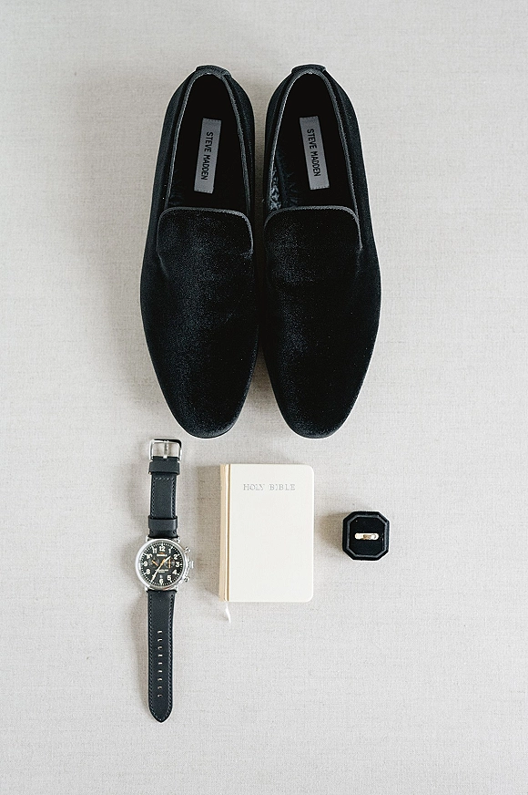 Groom accessories flat lay with black velvet loafers, wristwatch, Holy Bible, and ring box with wedding band on neutral fabric backdrop