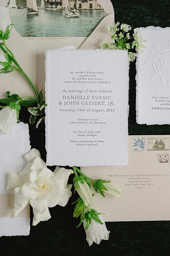 Invitation suite with deckle edge wedding invitation, embossed monogram cards, envelopes and stamps, styled with white roses on a dark tabletop