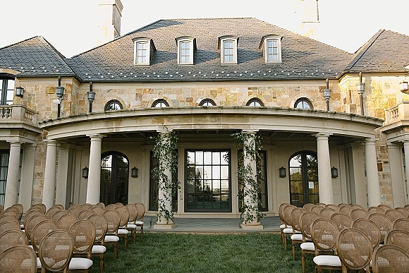 Ceremony setup with outdoor ceremony seating, rattan aisle chairs in neat rows on a lawn before a stone facade with arched windows and doors