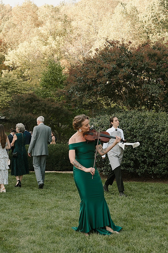 Wedding cocktail hour with a wedding violinist playing on a garden lawn as guests mingle; server in apron and green gown guest nearby