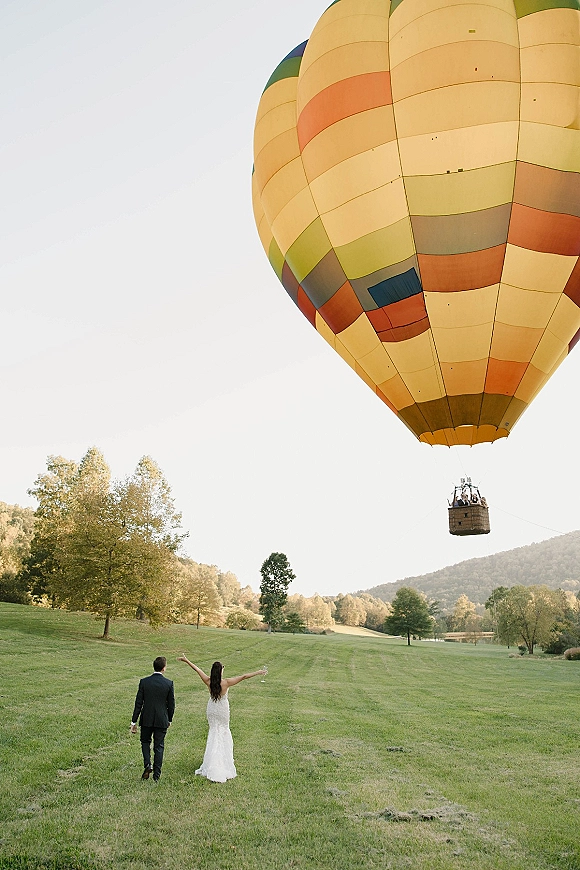 Couple portrait with a hot air balloon in the distance, bride in wedding dress and groom in suit walking away across a grassy field.