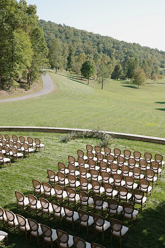 Outdoor ceremony setup with wood chairs and white cushions in a semicircle around low florals on a grassy lawn with rolling hills beyond