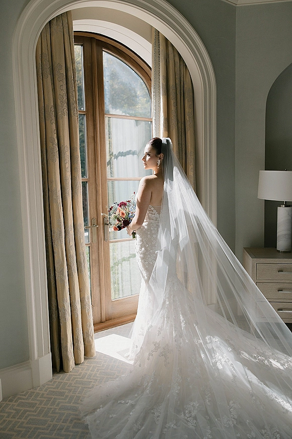 Bridal portrait of a bride by window holding a colorful bouquet, wearing a lace dress and long veil in a room with French doors
