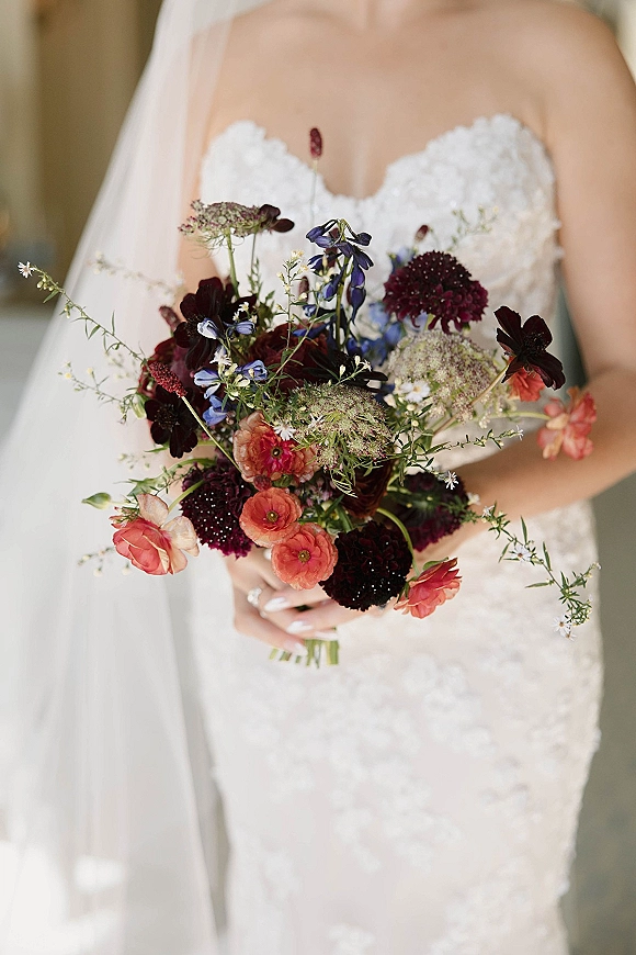 Bridal bouquet, wildflower bridal bouquet with coral, burgundy, and blue blooms held by a bride in a strapless lace gown and veil indoors