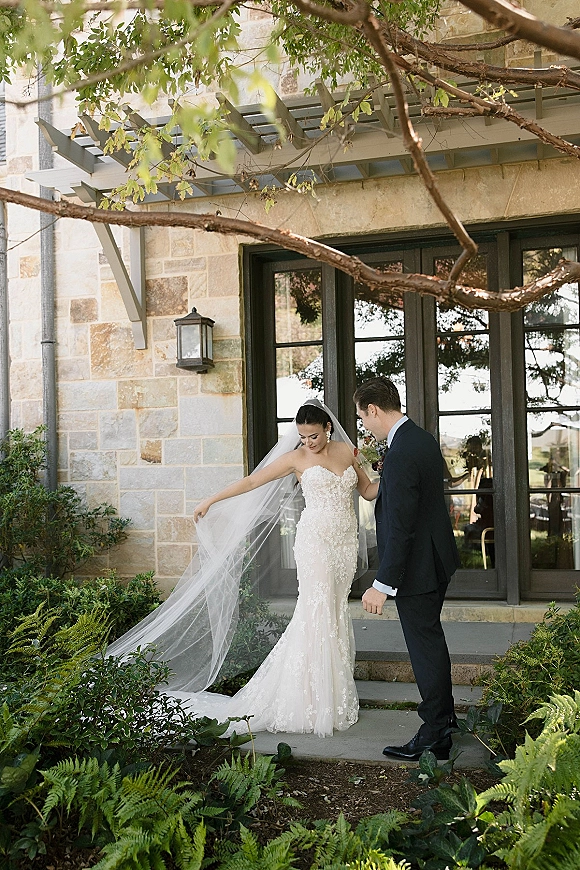 Couple portrait of bride and groom as she displays a cathedral veil and bouquet on stone steps by glass doors and greenery
