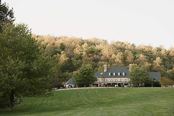Wedding venue exterior of a stone manor house with slate roof and chimneys, guests on the terrace overlooking rolling lawn and forested hills