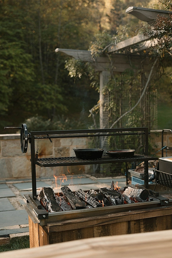 Outdoor grill setup with cast iron pots on a live fire cooking station, flames and smoke rising on a stone patio by garden trees