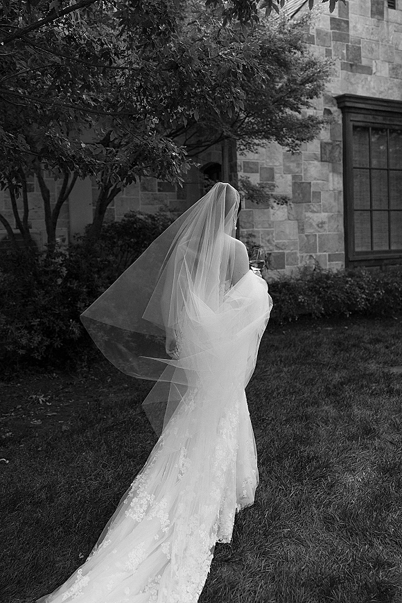 Bridal portrait in black and white of a bride from behind, long veil and lace dress train flowing as she holds a champagne glass by a stone building
