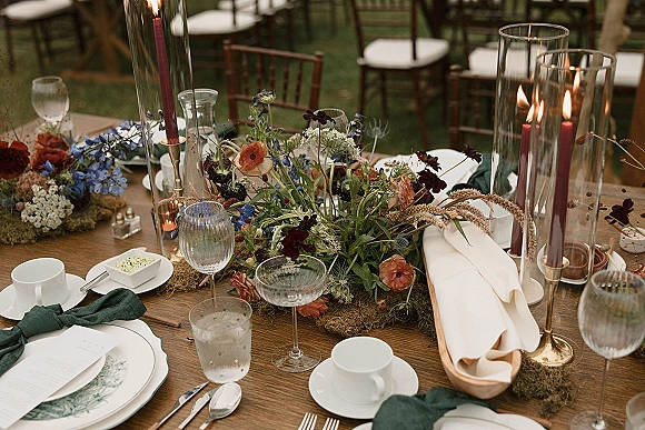 Reception tablescape with wildflower wedding centerpiece on a moss runner, taper candles in hurricanes, layered place settings on an outdoor lawn