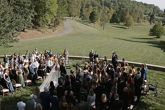 Wedding ceremony on a grassy lawn with guests standing in the round as the bride walks the aisle in a cathedral veil, hills behind