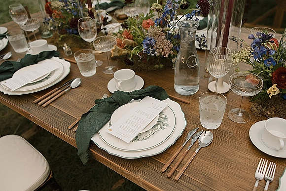 Reception tablescape on a wood wedding table with green linen napkins, patterned plates, mixed glassware, moss runner, florals, and taper candles on a lawn