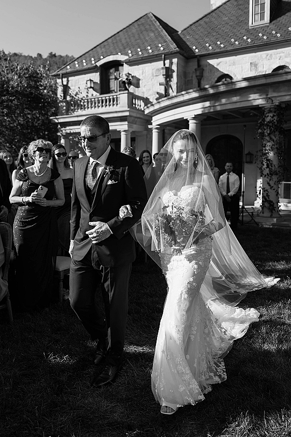 Wedding processional as bride walking down aisle with her father, holding a white and green bouquet, long veil, guests on a lawn by a stone estate