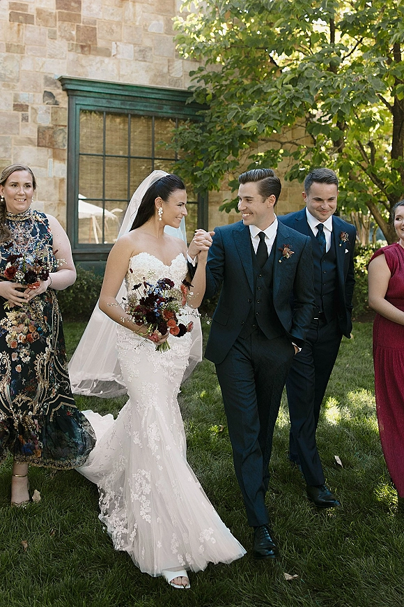 Wedding recessional with bride and groom walking hand in hand, smiling as she holds a bouquet, wedding party behind by a stone building
