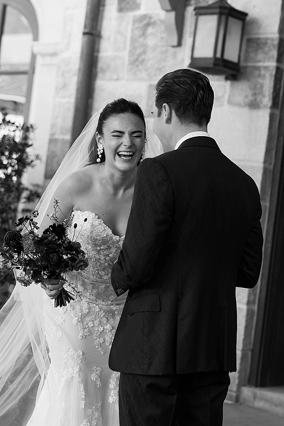 Couple portrait of bride laughing with groom, veil and bouquet visible, posed by a stone building’s arched doorway and lantern