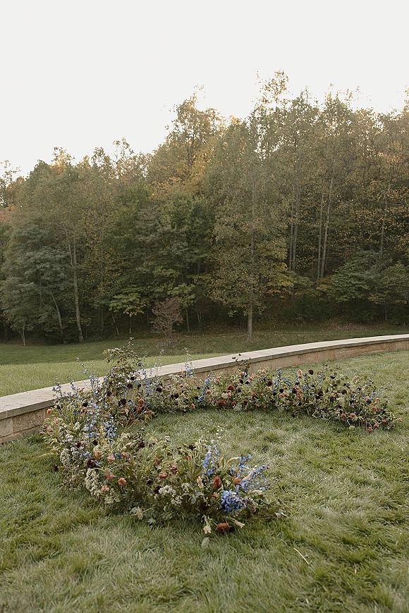 Ceremony aisle flowers with ground floral aisle wildflowers forming a low border along a grass lawn, set before a stone wall and forest hillside backdrop