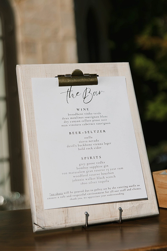 Wedding bar menu clipboard sign with black and white script on paper, clipped on a wood stand atop a tabletop with greenery blurred behind