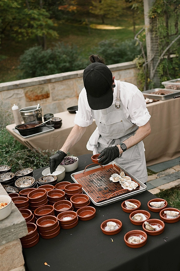 Wedding catering station with oysters on half shell arranged on trays as a chef in black gloves preps food beside chafing dishes in a garden patio setting