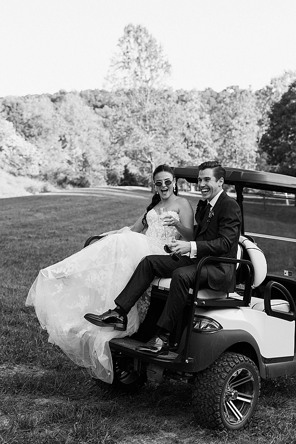 Wedding couple portrait of bride and groom on a golf cart laughing with champagne flutes, bride in sunglasses on a grassy lawn with trees