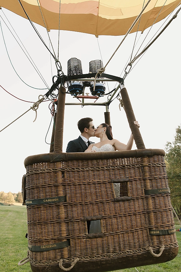Wedding kiss in a hot air balloon wedding basket, bride in veil and sunglasses with groom in tux, floating above a grassy field sky