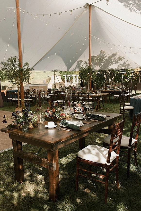 Reception tablescape for an outdoor tented reception on a dark wood farm table with floral centerpiece, taper candles, and string lights under a sail tent